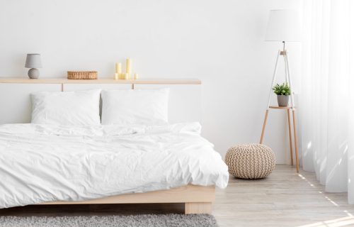 Double bed, lamp with plant in bedroom, soft gray carpet and ottoman, furniture with accessories Double bed, lamp with plant in bedroom, soft gray carpet and ottoman, furniture with accessories, window with curtains, white wall. Modern minimalist interior, scandinavian design, real estate blog ad